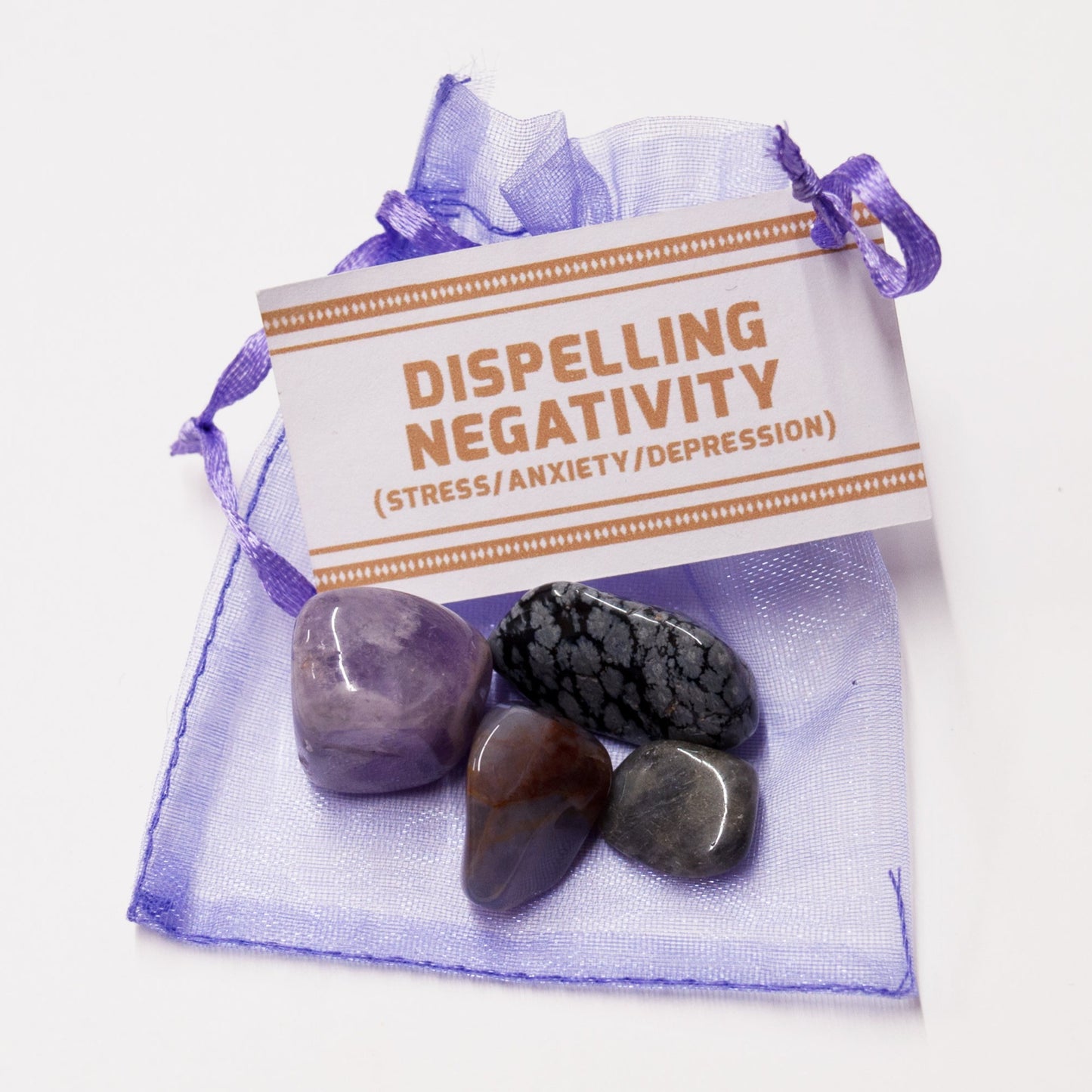 Four different tumbled crystals sit on a purple organza bag with a label stating Dispelling Negativity (Stress/Anxiety/Depression).